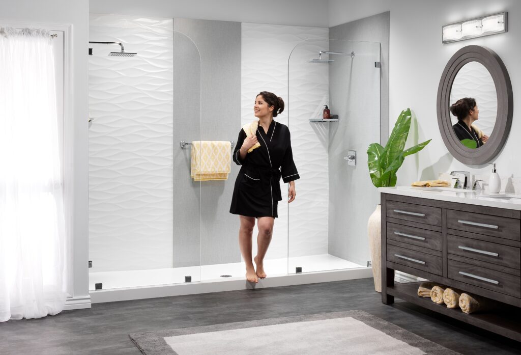 Woman in a black bathrobe standing inside a glass-walled shower, towel draped over her shoulder in a modern white-and-gray bathroom with a round framed mirror and vanity on the right.