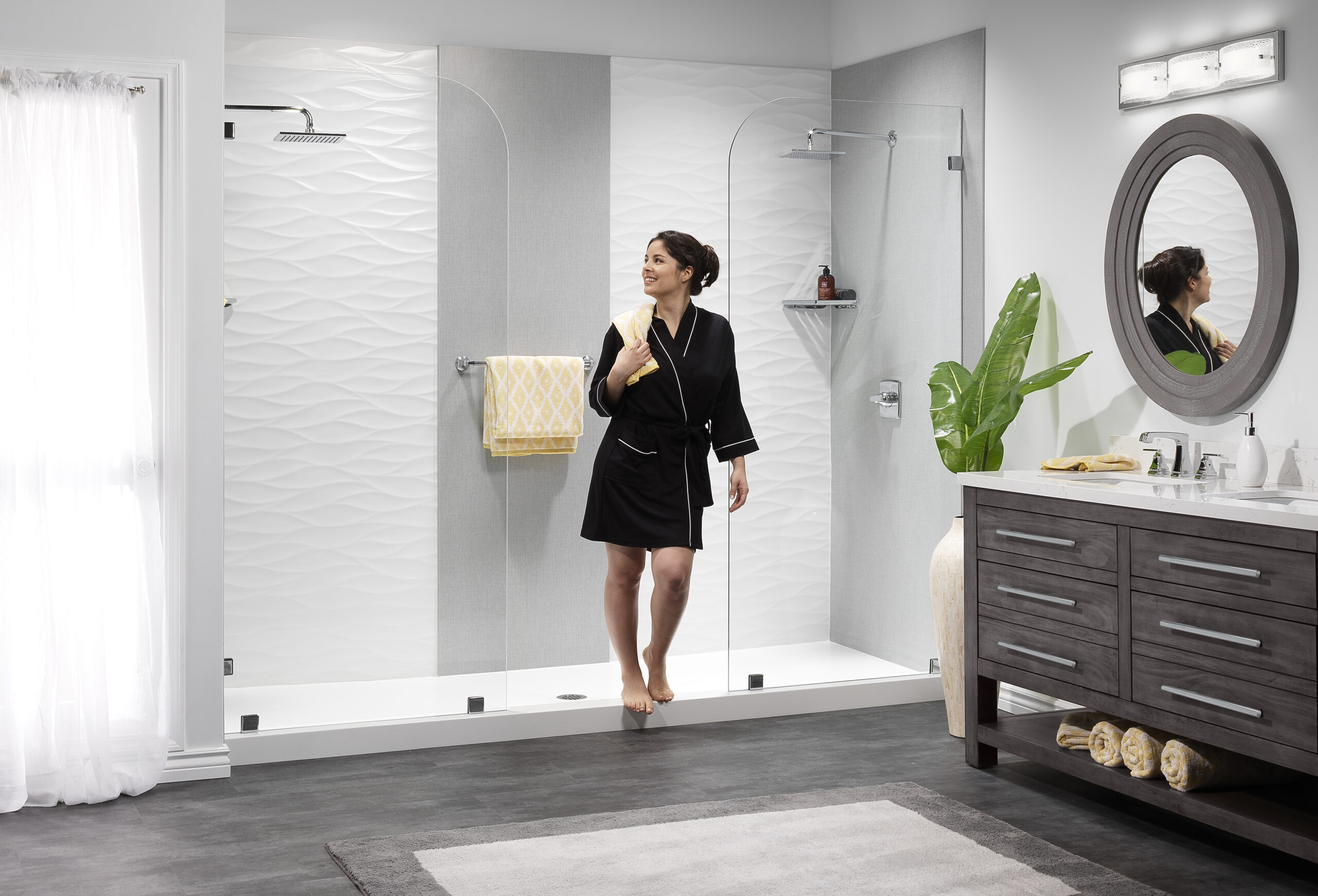 Woman in a black robe stands in a modern bathroom near a glass-enclosed shower, holding a yellow towel with a textured white wall behind and a plant beside a dark wood vanity.