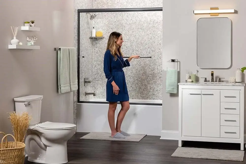 Woman in a dark blue robe standing on a bath mat in a modern bathroom, reaching toward the glass shower enclosure.