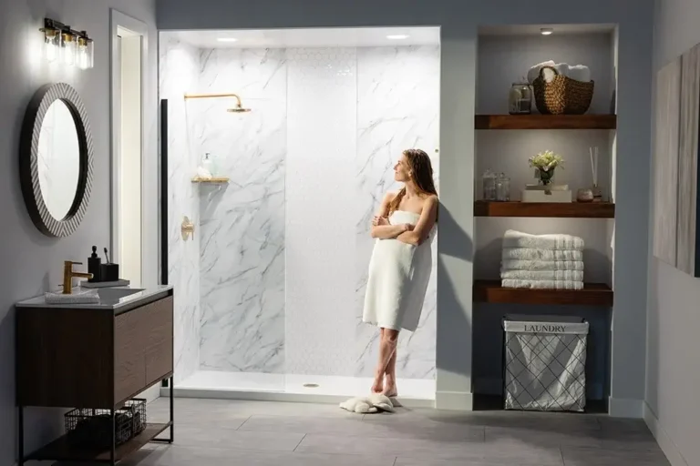 Woman in a white towel leans against the wall beside a glass-walled marble shower in a modern bathroom.